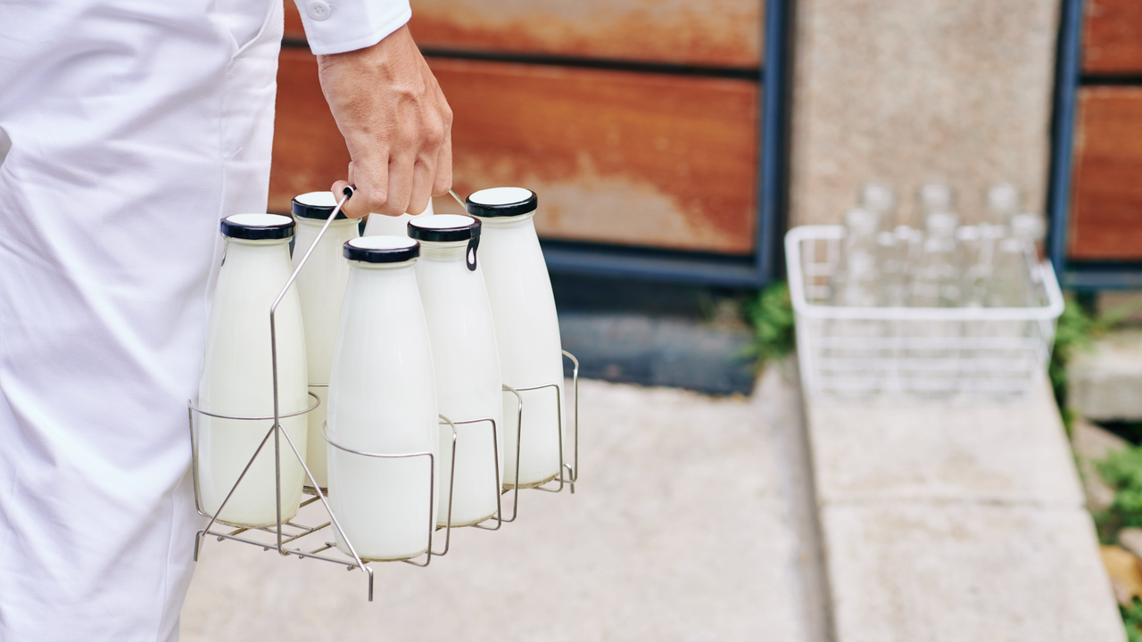 A milkman's arm holding a 6-pack of milk bottles as he walks towards a front door.